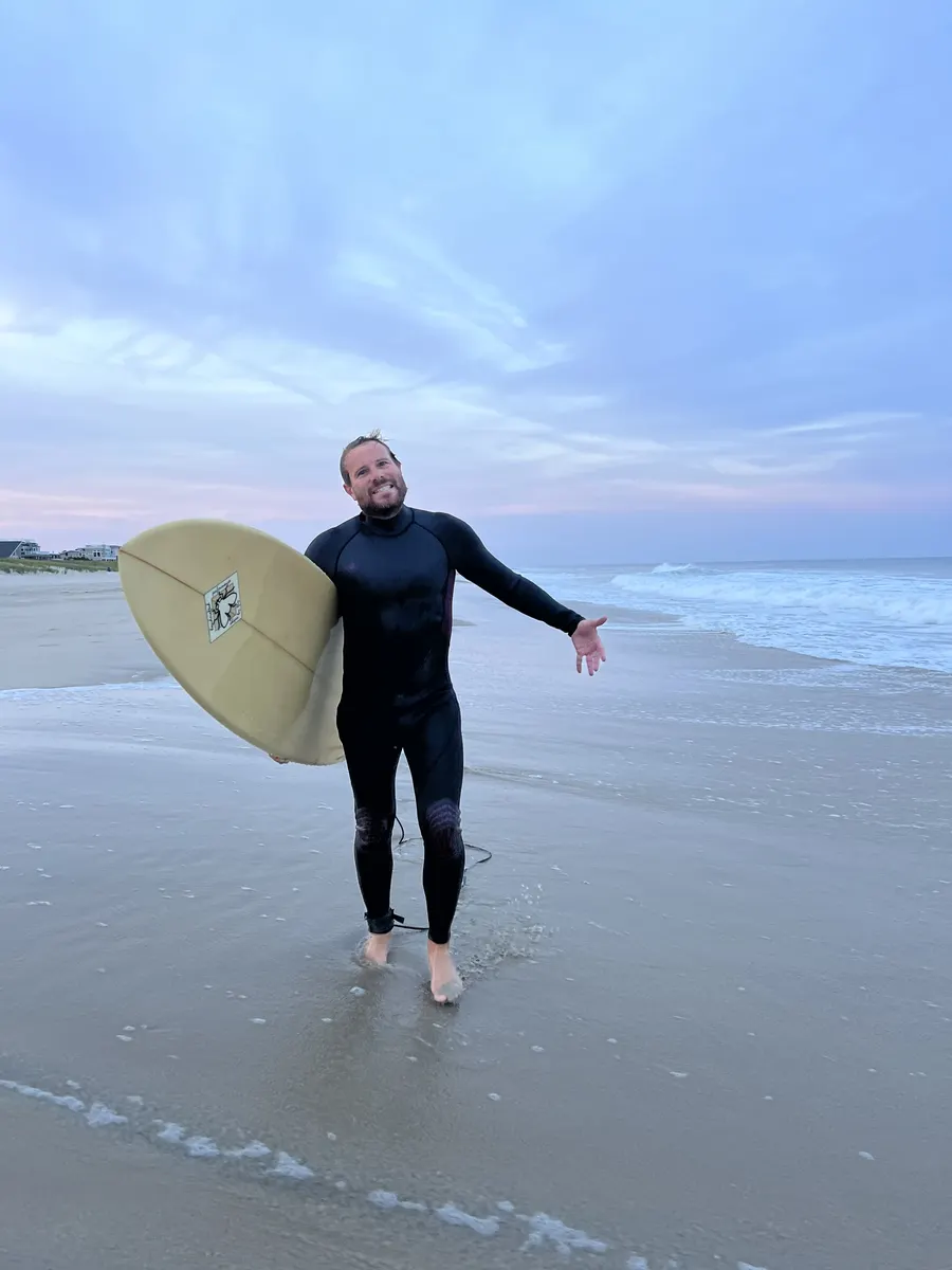 Brent in a wetsuit on the beach, holding a surfboard with arms wide at sunset