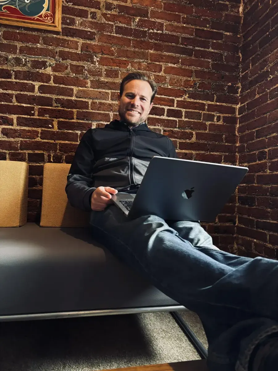 Brent Nickles relaxing on a leather couch with a laptop, in front of an exposed-brick wall
